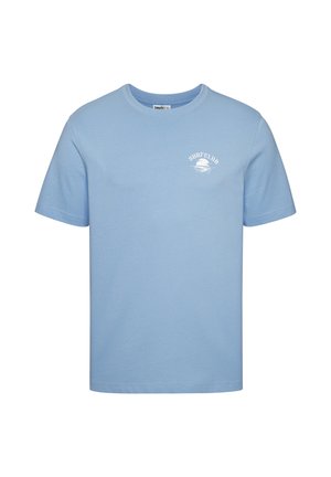 Hombre con camiseta azul clara con "SURF CLUB" blanco y gráfico de palmera, de pie junto a una pared blanca en una playa con el océano al fondo.