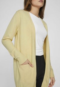 Ljusgul kofta med avslappnad passform, ribbmuddar, två framfickor och öppen framsida, lagd över en vit topp.