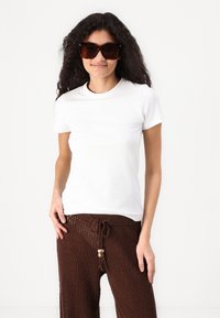 Camiseta blanca de manga corta, combinada con pantalones marrones texturizados que cuentan con un cordón, modelo usando gafas de sol oscuras de gran tamaño.