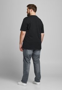 Schwarzes Baumwoll-T-Shirt mit kurzen Ärmeln, lockere Passform, kombiniert mit ausgewaschenen grauen Jeans und weißen Sneakers, von hinten betrachtet.