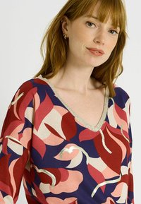 Suéter de manga larga con escote en V que presenta un patrón floral en rojo, rosa y azul marino, con acentos plateados a lo largo del escote y los contornos del diseño.