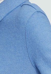 Gros plan de l'épaule d'une veste en tissu texturé bleu clair avec coutures visibles et matériau doux.
