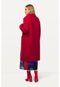 Manteau en laine rouge avec un large col, des manches longues et des poches latérales, associé à une jupe à motifs bleu et rouge et des bottes à talons rouges assorties.