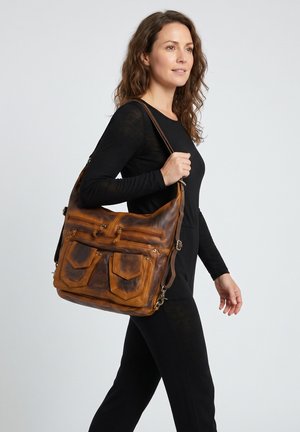 Vrouw met golvend haar in een zwarte outfit draagt een grote bruine leren tas met meerdere voorvakken over haar schouder.
