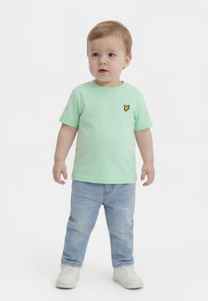 Dieťa s krátkymi svetlými vlasmi, oblečené v svetlozelenom tričku, svetlomodrých džínsoch a bielych teniskách, stojace pred jednoduchým bielym pozadím.