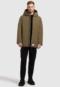 Olivgrön huva jacka med dragkedja fram, vadderad design och ett litet logomärke, i kombination med svarta cargobyxor och svarta stövlar.