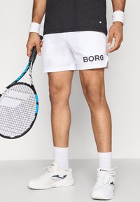 Calções de ténis brancos com o logo "BORG" em preto, combinados com sapatilhas brancas. Um esquema de ténis é segurado na mão direita.