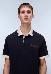 Μπλε ναυτικό πόλο πουκάμισο με μπεζ γιακά και μανσέτες, με μπορντό λογότυπο στο στήθος, τρία κουμπιά και υφή υφάσματος.