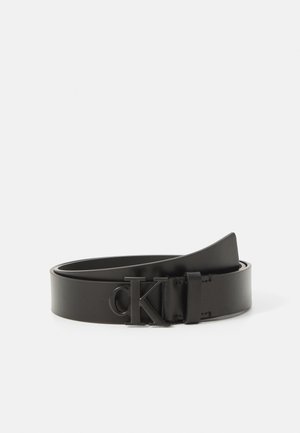 Zwarte leren riem met een matte zwarte gesp met het "cK" logo, opgerold op een witte achtergrond.