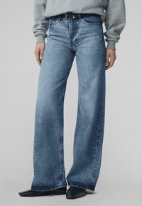 Asmuo dėvi šviesiai mėlynas plačias džinsines kelnes, juodą diržą, pilką džemperį ir juodus smailius batučius stovėdamas prie lygaus fono.