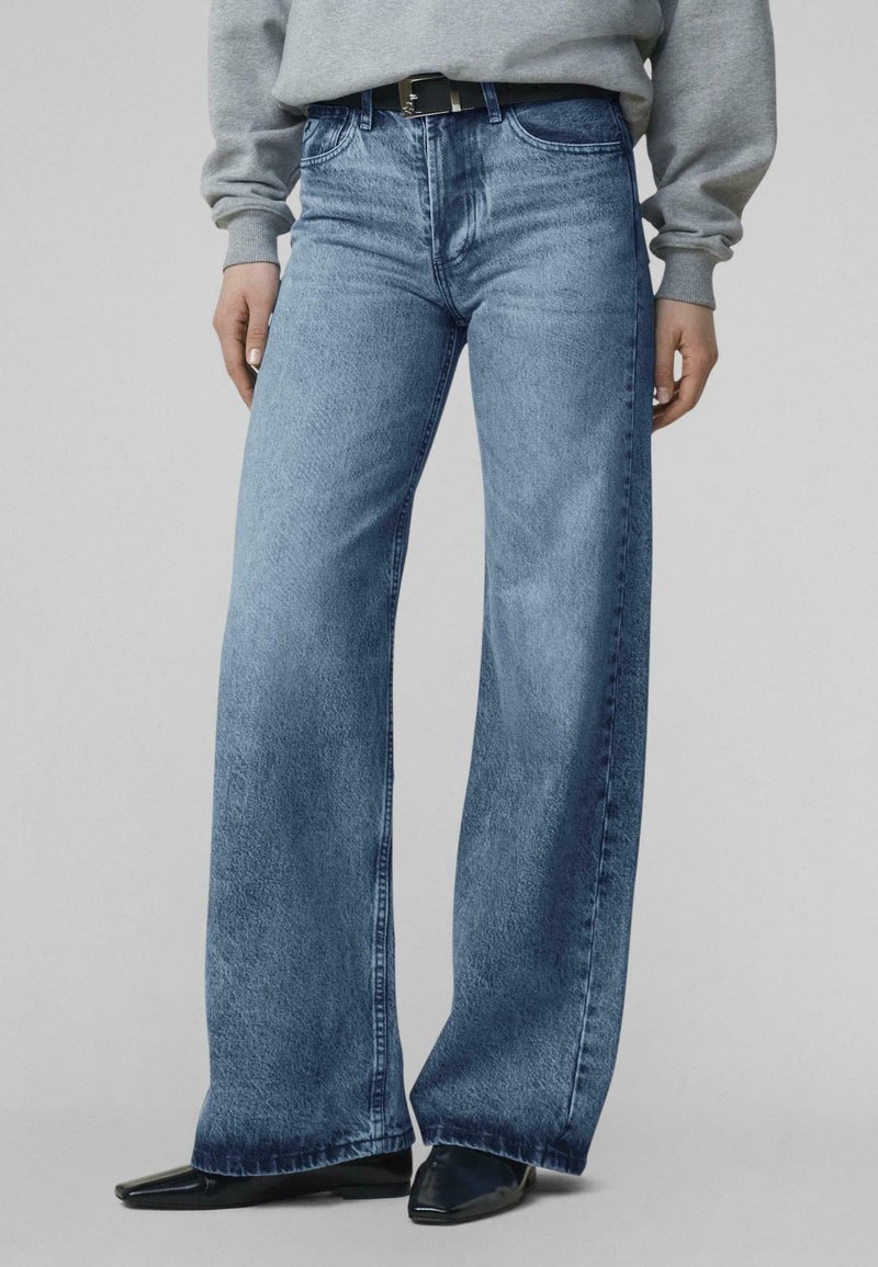 Asmuo dėvi šviesiai mėlynas plačias džinsines kelnes, juodą diržą, pilką džemperį ir juodus smailius batučius stovėdamas prie lygaus fono.