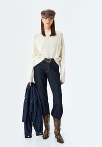 Maglione di maglia crema, pantaloni in denim scuri a vita alta, stivali da cowboy marroni con ricami floreali e un berretto di pelle marrone tenuto con una giacca.