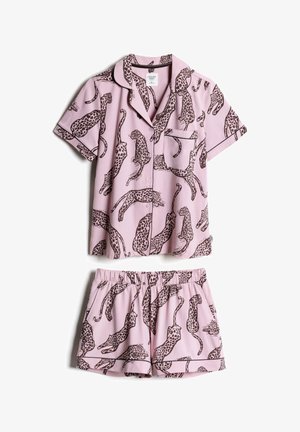 Junge Frau mit langen schwarzen Haaren, die ein pinkfarbenes kurzärmeliges Shirt mit schwarzem Leopardenmuster trägt und zur Seite schaut.