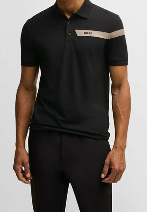 Mann trägt schwarzes kurzärmeliges Polohemd mit beigem Streifen über der Brust und „BOSS“-Logo, kombiniert mit schwarzen Hosen, stehend vor grauem Hintergrund.