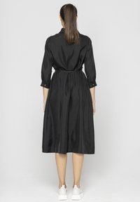 Czarna, długa sukienka koszulowa z paskiem w talii, do kolan, wykonana z lekkiego materiału z delikatnym połyskiem, z zapięciem na guziki przy mankietach, zestawiona z białymi trampkami.