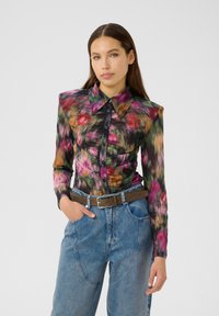 Frau mit langen braunen Haaren, die eine mehrfarbige Bluse mit Blumenmotiven und Puffärmeln sowie hochtaillierte blaue Jeans mit einem braunen Gürtel trägt.