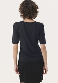 Person mit lockigem braunem Haar, der ein schwarzes Kurzarmoberteil und einen schwarzen Rock trägt und mit dem Gesicht von der Kamera weg vor einem schlichten Hintergrund steht.