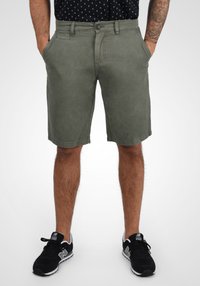 Shorts de culoare verde măsliniu, dintr-un material moale, cu talie standard și buzunare frontale, asortați cu adidași negri.