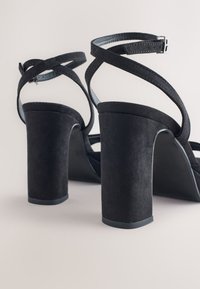 Sorte ruskinds-sandaletter med høje hæle og blokhæl. Har krydsede ankelstropper og en glat inderforing. Tekstureret overflade.