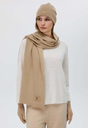 Cappello e sciarpa a coste beige in materiale morbido, abbinati a un maglione bianco soffice. Entraccessori presentano un discreto marchio.