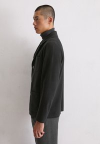 Schwarzer Wollblazer mit tailliertem Schnitt, einer aufgesetzten Tasche und Knopfverschluss. Tragen über einem dunklen Rollkragenpullover, im Profil vor hellem Hintergrund dargestellt.