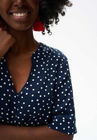 Marineblouse met witte stippen, V-hals, korte mouwen en een gladde stof. Rode kwastoorbellen voegen kleurcontrast toe.