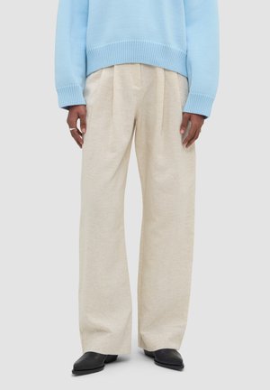 Person trägt lockere beige Hose, schwarze spitze Schuhe und einen hellblauen Strickpullover, steht vor einem schlichten Hintergrund.