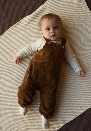 Pruunid sametpüksid lillemustritega, kantud beežide pika varrukaga särgi peal, tekstuurse kreemja pleedi peal. Laps lebab ja vaatab üles.