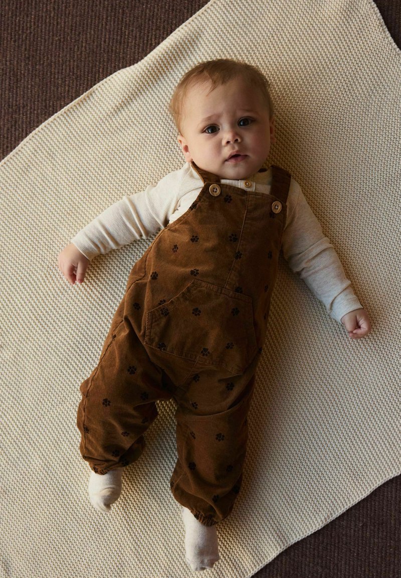 Salopette en velours côtelé marron avec des motifs floraux, portée sur un t-shirt à manches longues beige, sur une couverture crème texturée. L'enfant est allongé, regardant vers le haut.