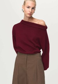Burgundowy sweter bez ramion wykonany z miękkiej dzianiny, zestawiony z brązowymi spodniami z wysokim stanem, które mają plisowania i gładką fakturę.