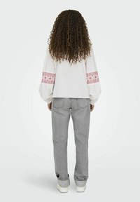 Balta blusa su raudonais siuviniais ant rankovių, derinama su šviesiai pilkomis džinsais. Modelis stovi nugara į kamerą, matyti garbanoti plaukai.