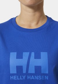 Modrá bavlněná tričko s velkým světle modrým logem "HH" a textem "Helly Hansen" na spodní části. Kulatý výstřih s hladkým povrchem.