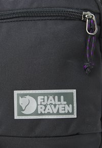 Zaino in tessuto nero con un prominente patch del logo Fjällräven. Presenta una tasca con zip e un cordino arricchito da dettagli viola. Superficie texturizzata.