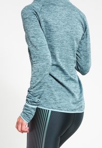 Langärmliges Sportoberteil in meliertem Petrol mit strukturiertem Stoff und Daumenlöchern, kombiniert mit schwarzen Leggings mit vertikalen Streifen.