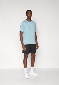 Αθλητική μπλούζα σε ανοιχτό γαλάζιο χρώμα με κοντά μανίκια και λευκό λογότυπο, σε συνδυασμό με μαύρο σορτς και λευκά αθλητικά παπούτσια. Τοποθετημένα σε ουδέτερο φόντο.