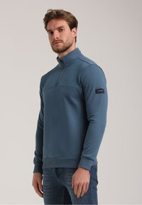 Sinine pullover-sweater, millel on pooltõmbe zipp ees ja tekstuuriga paneel rinna piirkonnas. Omab soonikuid mansette ja logo paika varrukal.