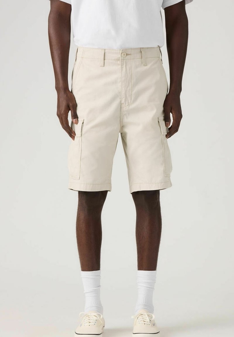 Homme portant un short cargo beige, des chaussettes blanches et des baskets beiges, debout devant un fond uni.