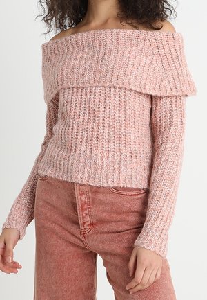 Vrouw die een roze gebreide trui met blote schouders draagt en een roze high-waisted jeans, staand tegen een effen lichte achtergrond.