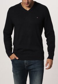 Pull noir à col en V en tissu tricoté doux, avec poignets et ourlet côtelés, orné d'un petit logo sur la poitrine. Porté avec un jean bleu.