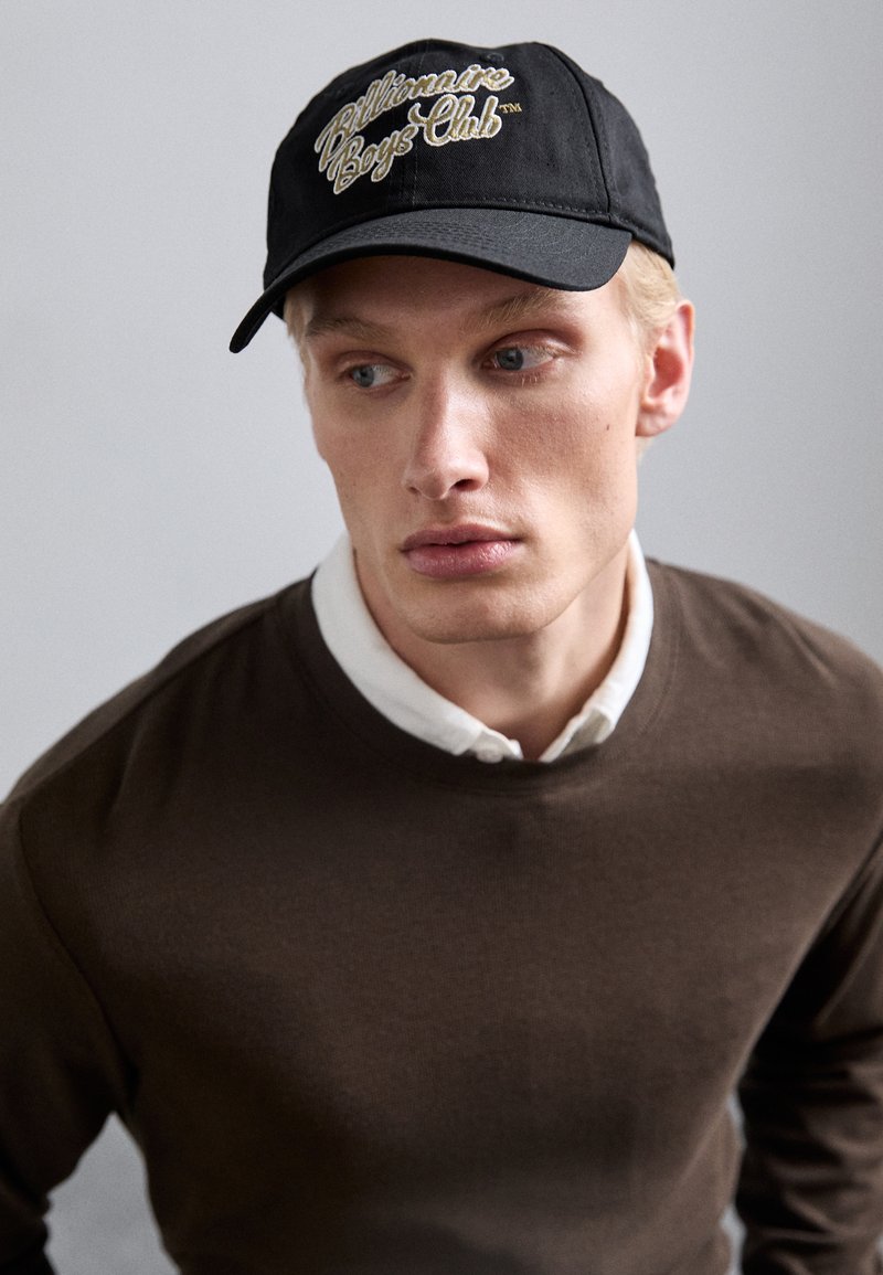 Musta baseball-lippis, johon on brodeerattu "Billionaire Boys Club" -teksti, yhdistettynä ruskeaan huppariin ja valkoiseen kauluspaitaan.