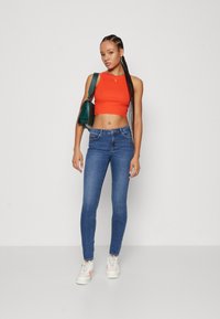 Red cropped tank top, blue skinny jeans, and white sneakers with pastel accents. Model holds a green bag and has braided hair.