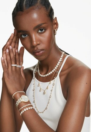 Ung kvinde med flettet hår iført lagdelte guld- og diamantjuveler, herunder halskæder, armbånd, ringe og et ur, iført en hvid top.