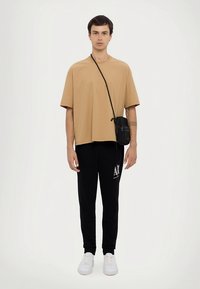 Tan oversized t-shirt with short sleeves, paired with black joggers featuring a white logo, and completed with white sneakers and a small black bag.