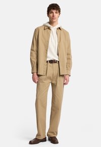 Camicia button-up beige chiaro sopra un felpa con cappuccio bianca, abbinata a pantaloni beige e scarpe di pelle marroni. Texture liscia su tutta la superficie.