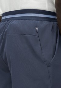 Gros plan d'une personne portant un pantalon bleu marine avec une taille élastique comportant une bande bleu clair et une petite poche zippée.