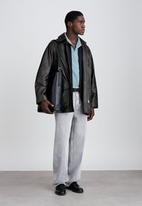 Melnas ādas jaka, zila polo krekls, pelēki plati bikses un melnas kurpes. Cilvēks tur melnu somu ar siksnu.