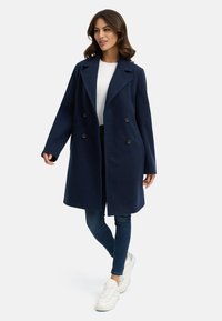 Mörkblå ullkappa med dubbelknäppt design, slagkrage och två framfickor, bärs över en vit skjorta och blåjeans.