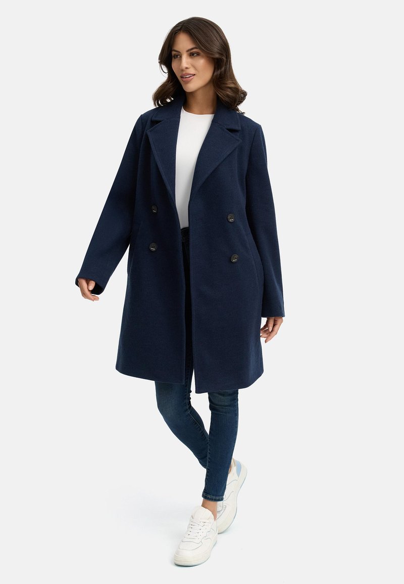 Mörkblå ullkappa med dubbelknäppt design, slagkrage och två framfickor, bärs över en vit skjorta och blåjeans.
