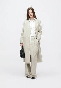 Femeie purtând pantaloni bej cu picioare largi, palton lung asortat, cămașă albă, pantofi negri, ținând o geantă mică neagră, stând pe fundal alb.