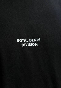Schwarzer Stoff mit weißem Text "ROYAL DENIM DIVISION" in Großbuchstaben in der Mitte gedruckt.